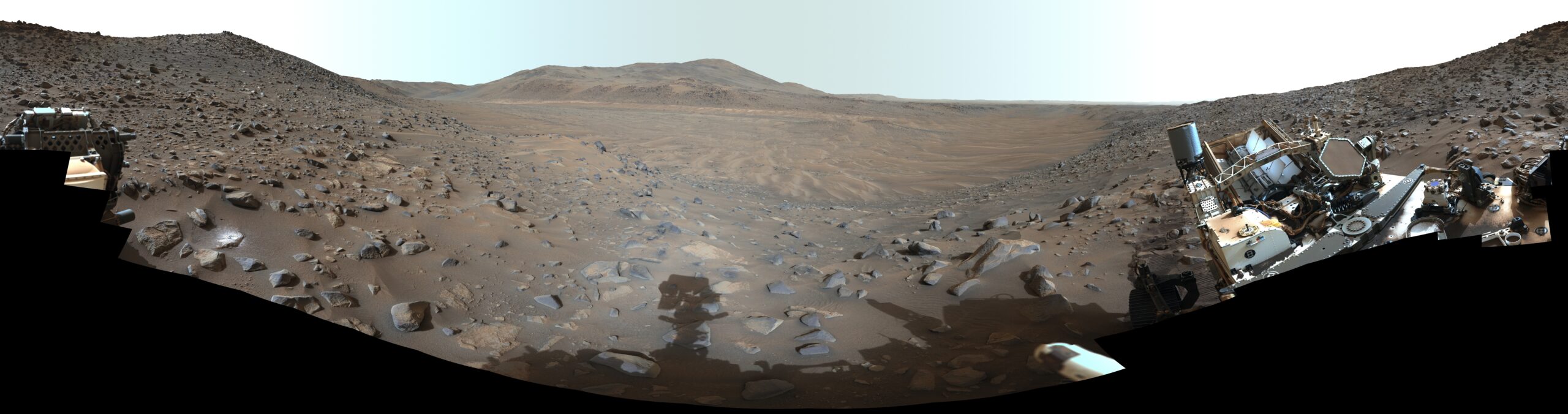 A panoramic view of a barren, rocky landscape with distant hills, framed by parts of a robotic ro...