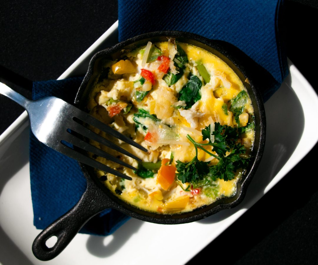 A cooked egg frittata with colorful vegetables sits in a small black cast iron and serving dish a...