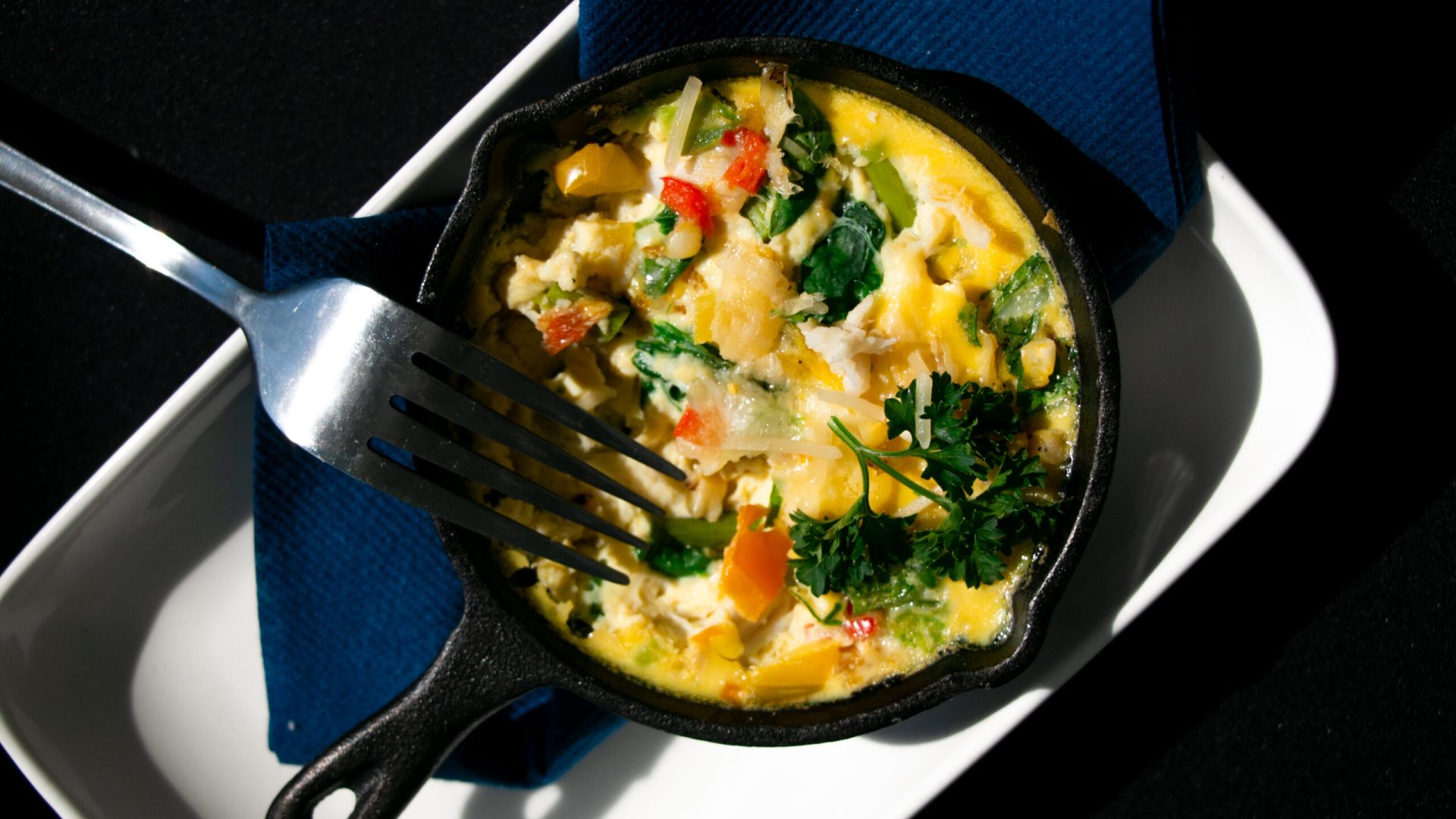 A cooked egg frittata with colorful vegetables sits in a small black cast iron and serving dish a...